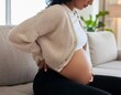 © IWei - A black pregnant woman sitting on a couch indoors, with one hand on her lower back and the other resting on her bare baby bump
