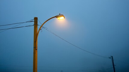 Poster - Soft yellow light from a vintage streetlight breaks through a misty blue-gray background, enhancing the serene atmosphere of the urban landscape during twilight