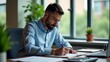 © Zaahid - Focused man in a blue shirt writing at a desk with natural light from the window illuminating the office space
