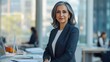 © Daiki - A confident mature Middle Eastern woman in her 50s stands in a modern office environment, exuding professionalism and elegance. Her stylish appearance reflects leadership qualities.