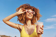 © SHOTPRIME STUDIO - Happy woman with curly hair wearing sunglasses and holding a milkshake against a blue sky
