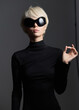 © SHOTPRIME STUDIO - Stylish woman with short blonde hair wearing oversized black sunglasses and a form-fitting black turtleneck, posing confidently against a dark background.