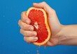 © Ozz - Hand squeezing a grapefruit half, creating fresh juice against a bright blue background.