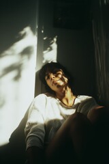 young woman sits quietly, sunlight illuminating her face and casting leaf shadows on the wall behind