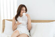 © Rakchanok - young, woman, beauty, lifestyle, coffee: Asian woman in white shorts savors warm drink on bright bed, smiling, radiating glow and calm routine that pairs self-care with easy morning comfort, holiday