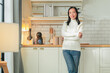 © whyframeshot - Portrait of a Asian smiling mature woman casual cloth standing in her kitchen confident cheerful looking at camera woman in white sweater standing in kitchen daylight