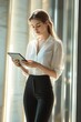 © Jdgkxbnc - Young woman standing indoors near a window looking thoughtfully at a tablet device in her hands with soft natural lighting
