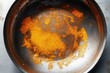 © zong - Close-up of a stained and burnt metal frying pan with orange and brown residue inside, showing signs of heavy use and burning