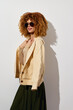 © SHOTPRIME STUDIO - Fashionable woman with curly hair wearing sunglasses and a beige cardigan, standing against a white background, showcasing a stylish combination of casual wear
