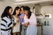 © Drobot Dean - Four young women stand together in a modern kitchen, laughing and smiling while looking at a smartphone. They are enjoying a casual moment indoors during the daytime.