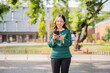 © maeching - Asian female student in warm clothes using phone to chat with friends and ready for video call while in university.