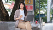 © wattana - Woman in smart casual outfit sitting on a curved bench, multitasking with coffee and laptop.