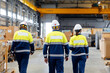 © Parilov - Group of warehouse workers wearing hard hats and reflective jackets uniform waking in industrial hangar of factory, back view