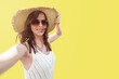© Vladimir Razgulyaev - A smiling woman of 30-35 years old in a wide-brimmed hat and sunglasses on a yellow background, she takes a selfie. A concept for travel and blogging