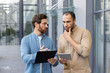 © Liubomir - Two men are engaged in a discussion outdoors. One holds a clipboard, while the other considers a tablet in their hands.