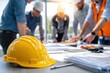 © Denis Tuev - Yellow hard hat placed on a table with construction plans, while a group of professionals collaborates in the background, showcasing teamwork and project planning in a bright workspace