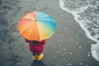 © Iryna - At the beach, a boy in a red raincoat and yellow rubber boots holds a rainbow umbrella, and a child with a colourful umbrella is looking out to sea, while a school kid wears a waterproof red coat and