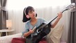 © FAMILY STOCK - Asian girl wearing headphones learning to play guitar at home using smartphone, showing modern self-learning and music practice in a cozy bedroom.