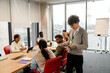 © Drobot Dean - A young White man presenting documents to a multinational group of people during an internship session inside a bright modern office with large windows, red chairs, and a wooden conference table.