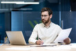 © Tetiana - Concentrated male banker and office worker sitting in the office at the table, working on a laptop and with documents