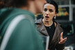 © qunica.com - A woman wearing a leather jacket gestures emphatically, showing a frustrated facial expression, while speaking to another person outdoors.