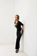 © Budi - Elegant woman in stylish black jumpsuit leaning against a white wall in studio