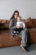 © Alex - Stylish young mother with long dark hair in a gray stylish suit with her son in her arms sitting on the sofa. Handsome son with mom