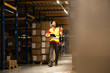 © Dejan - A warehouse worker with a tablet smiling while walking through an organized aisle filled with stacked boxes.