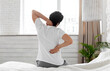 © Prostock-studio - Unrecognizable guy suffering from back pain in bedroom, waking up in morning, sitting on bed and touching his neck and lower back with both hands, wearing pajamas, copy space, back view