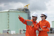 © eakgrungenerd - Professional engineer wearing orange suit and white hard hat stands using tablet in liquefied natural gas facility industrial structures petroleum. Survey planning for gas power transmission projects.