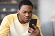 © Prostock-studio - African american guy squinting while looking at mobile phone screen at home, copy space, blurred background. Young black man reading suspicious message on his smartphone, closeup
