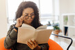 © Prostock-studio - Focused confused young black lady trying to read paper book, squinting to see more clearly, wearing glasses, having difficulties seeing text because of vision problems, sitting on pouf in living room