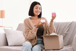 © Prostock-studio - Portrait of smiling asian lady received package, unpacking cardboard beauty box with cosmetics product, holding skincare cream in both hands sitting on the couch. Woman satisfied with online shopping