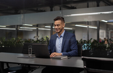 Positive young arabic manager working on laptop at modern hi-tech office, sitting at table, typing on computer keyboard, sending emails, communicating with business partners, copy space