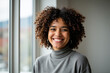 © Creuxnoir - Woman with curly hair smiling at camera in front of a window, with a wall in background.