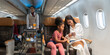 © boonsom - Woman in white outfit and young girl in pink clothing enjoying on mobile phone together in airplane cabin. Female flight attendant pushing service cart down aisle to passengers