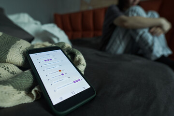 Image depicting a smartphone on a bed displaying a messaging app, with a person sitting in background appearing distressed and distant