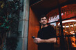© BullRun - Stylish young man with mobile phone in hand smiling subtly, standing near cozy restaurant entrance with warm lights and greenery, casual evening moment in urban setting.Young male using smartphone app