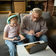 © master1305 - Elderly man and girl wearing headphones, using tablets and writing notes during online learning in cozy living room. Concept of elderly lifestyle, education, knowledge, childhood, development