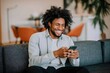 © Andrii Zastrozhnov - Smiling african male adult texting on smartphone in modern living room