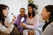 © Drobot Dean - Group of four female friends sitting on a bed and smiling while talking and applying makeup, in a bedroom