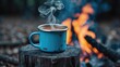 © TheWaterMeloonProjec - A blue enamel mug filled with hot coffee rests on a weathered log next to a campfire outdoors. Selective focus on the cup with a soft background.