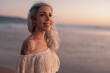 © kegfire - A confident mature woman with gray hair smiles serenely as she stands on the beach, enjoying a beautiful sunset. Her off-shoulder dress and visible tattoo add to her unique style.