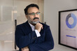 © fizkes - Thoughtful middle-aged Indian businessman dressed in suit standing in modern office next to screen displaying charts, focused on analytics, strategy planning and decision-making. Business intelligence