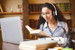 © Prostock-studio - Portrait of calm young woman wearing wireless headset, reading paper book, sitting at table with laptop computer. Lady studying online at home, doing homework, enjoying story, preparing for exam