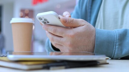 Person is using a cell phone while sitting at a table with a cup of coffee. The table is cluttered with various items, including a laptop, a book, and a few pens