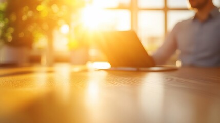 Aufkleber - Blurred image of a person working on a laptop at a wooden desk in a sunlit office.