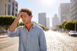 © Slepitssskaya - Young man in casual attire stands on a sunlit street, shielding his eyes from the intense heat, with shimmering pavement and urban buildings creating a vibrant summer atmosphere