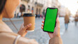 © anandart - Close-up of young casual woman drinking coffee holding vertical smartphone with green screen, sunny street, lifestyle and technology connection