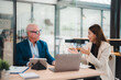 © Tj - Senior businessman and young businesswoman discussing strategy during a meeting in a modern office, using laptop and tablet, collaborating on a project and sharing ideas
